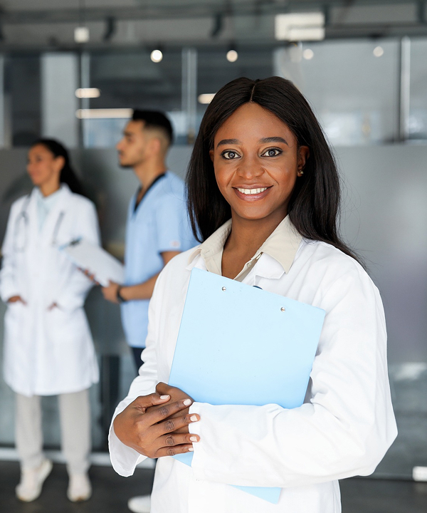 A smiling medical professional in a white coat holds a clipboard, with two other healthcare workers in the background.