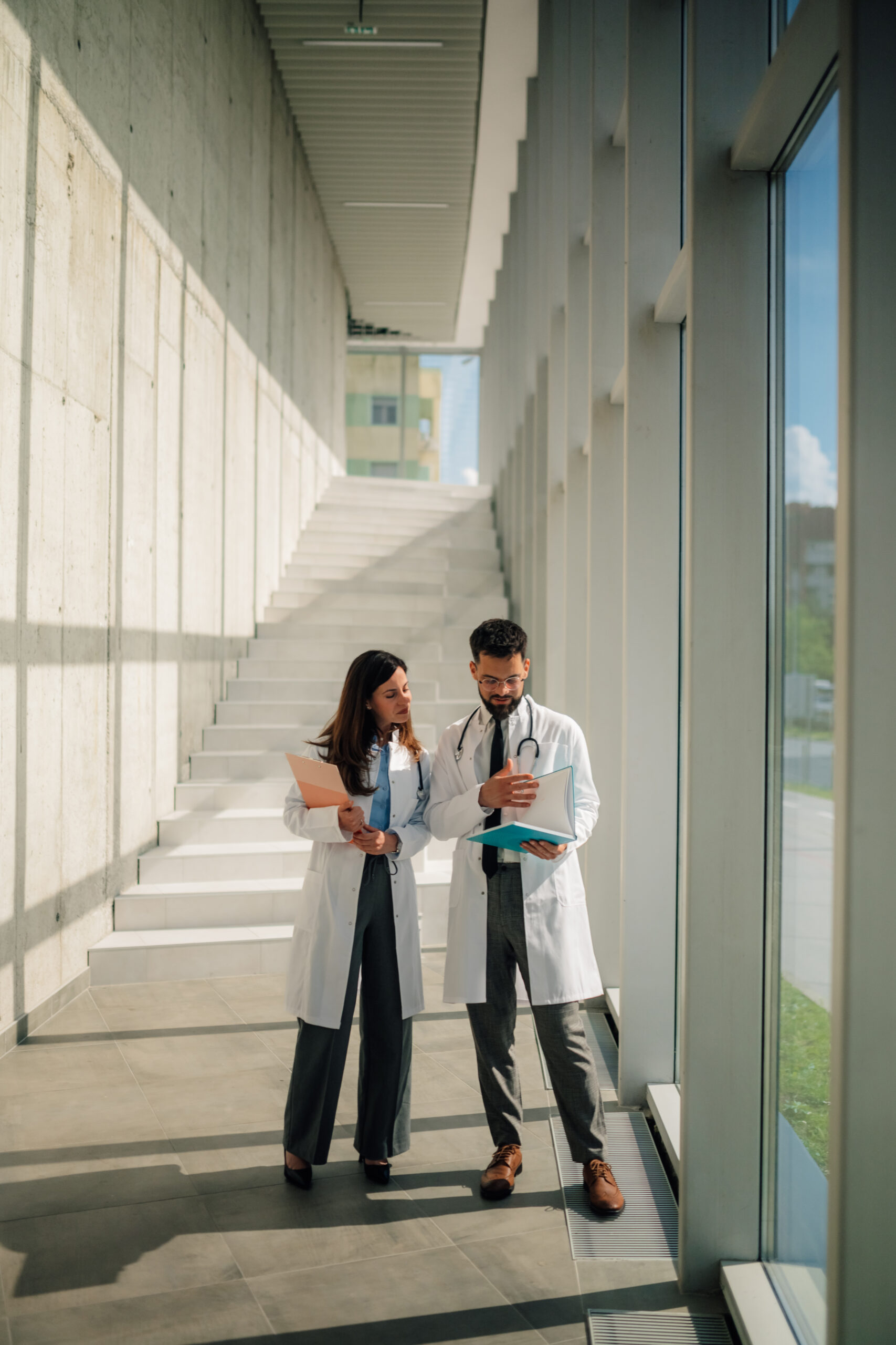 Two medical professionals discuss documents in a brightly lit, modern hallway.