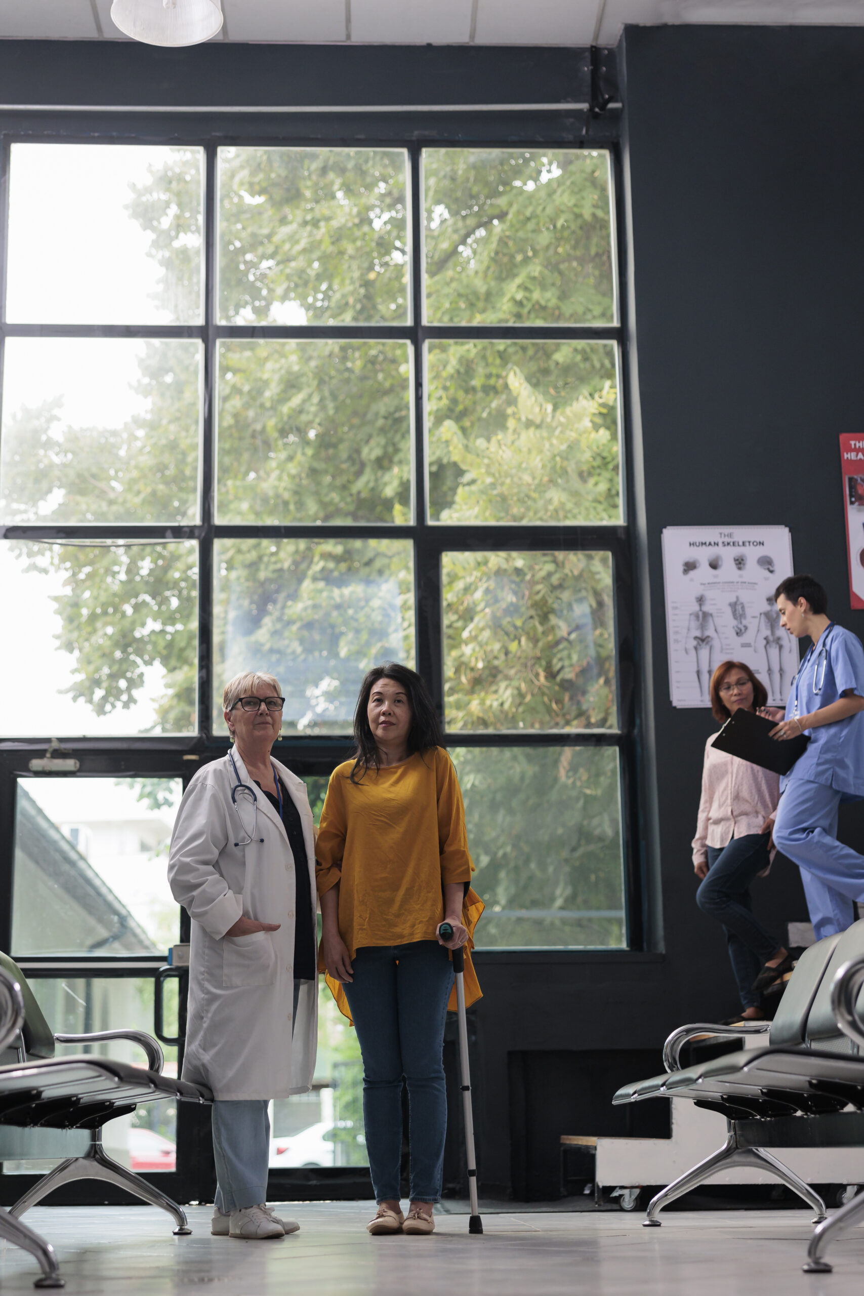 A doctor stands with a patient using a crutch in a medical setting, while others are in the background.